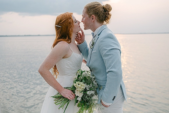 Couple portrait of bride holding a white-green bouquet and groom in a light blue suit, nose to nose by a lakeshore at sunset