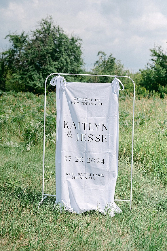 Wedding welcome sign, fabric wedding welcome sign on a metal frame with ribbon bows and modern typography in a tall grass meadow under cloudy sky
