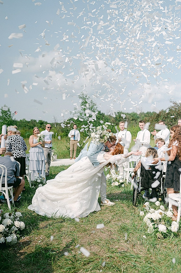 Wedding kiss moment with confetti wedding exit as the bride and groom dip kiss down a floral-lined aisle in a grassy field with guests cheering