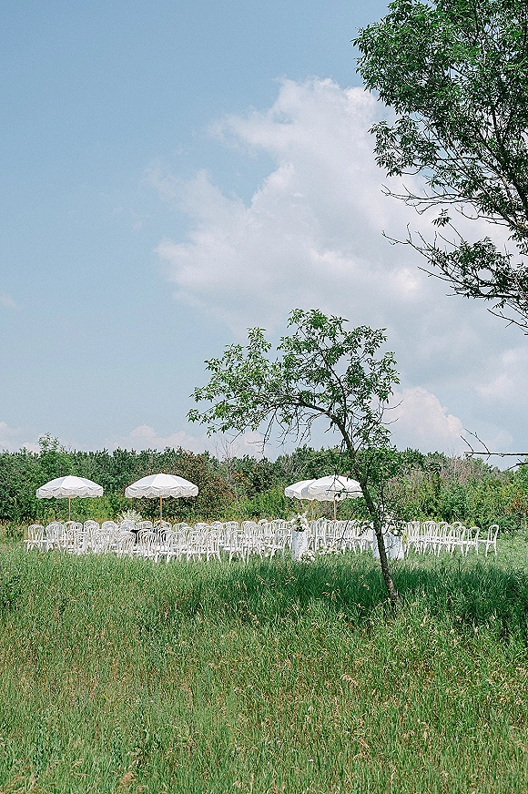 Ceremony seating with outdoor ceremony chairs arranged in rows, white umbrellas and draping in a meadow under blue sky and trees