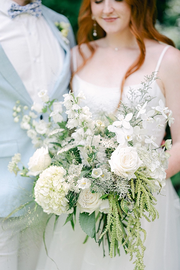 Bride holding bouquet of white roses, orchids and hydrangea with trailing greenery, groom in light suit beside her outdoors
