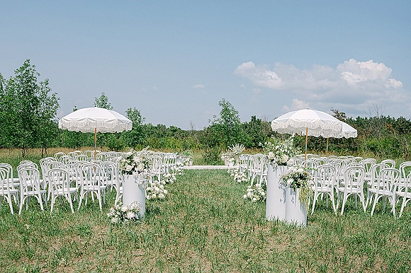 Outdoor ceremony setup with garden ceremony chairs and a white aisle runner, parasols and floral pedestals on a grassy field under blue sky