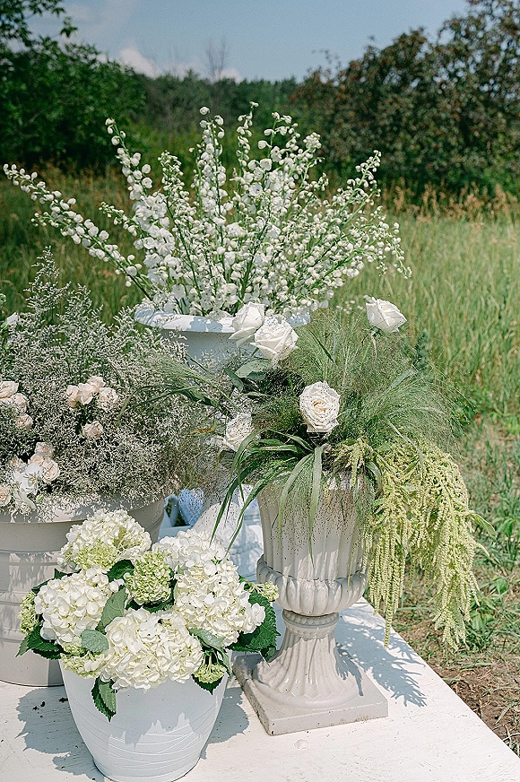 Wedding floral arrangements with white wedding flowers in ceramic buckets and a stone urn on a table, set in a meadow under blue sky