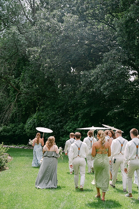 Wedding party walking with bridesmaids and groomsmen from behind, bouquets and parasols in hand along a grassy riverside lawn with trees