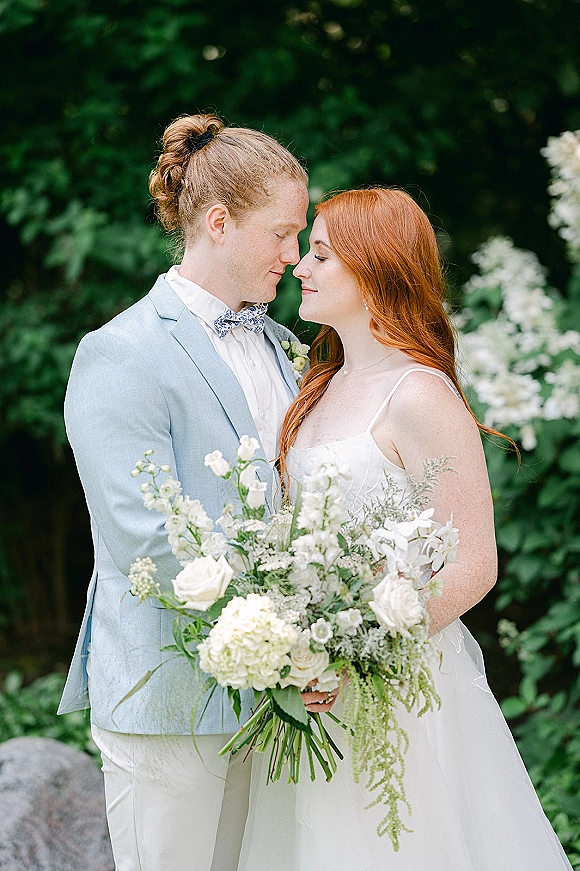 Couple portrait of bride and groom in a close embrace, touching foreheads as she holds a bridal bouquet in a garden setting