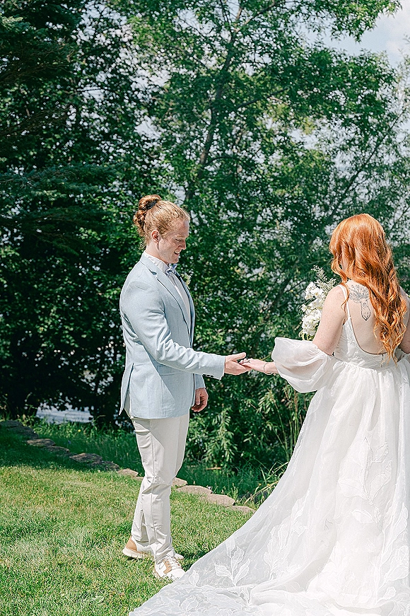 First look moment outdoors as bride in a puff sleeve wedding dress holds a white bouquet while groom in a light blue suit holds hands by a pond