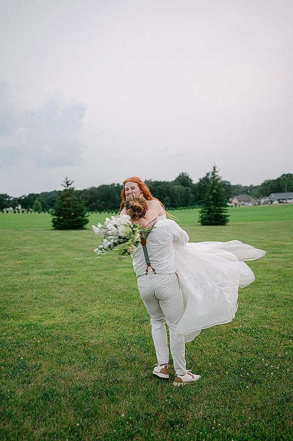 Couple portrait of bride and groom hug as he lifts her in a grassy field, her wedding dress train flowing under a cloudy sky