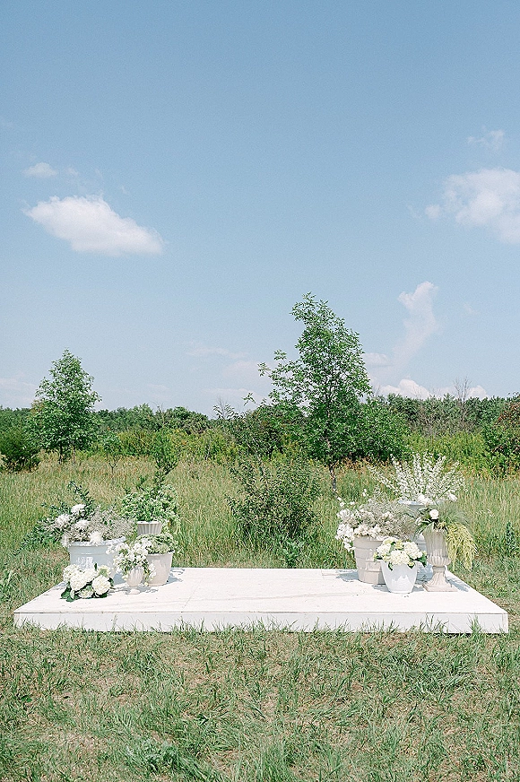 Ceremony backdrop decor with a white platform, asymmetrical white flowers and greenery in pedestal urns, set in a meadow field under blue sky