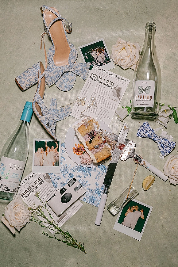 Wedding flat lay with wedding detail flatlay of blue floral bow heels, rings, roses, champagne, polaroids, and cake slice on neutral fabric
