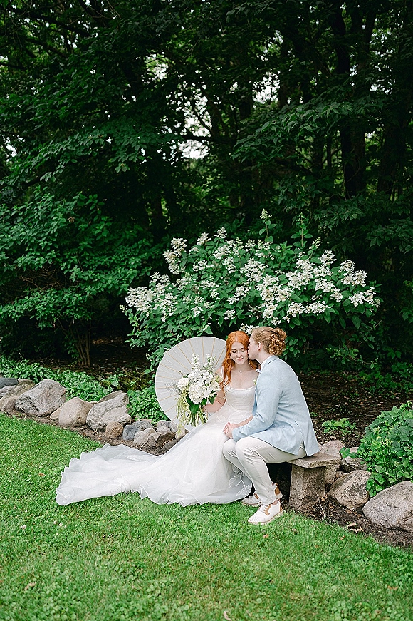 Couple portrait of bride and groom sitting on a garden bench, sharing a forehead kiss under a white parasol with bouquet nearby