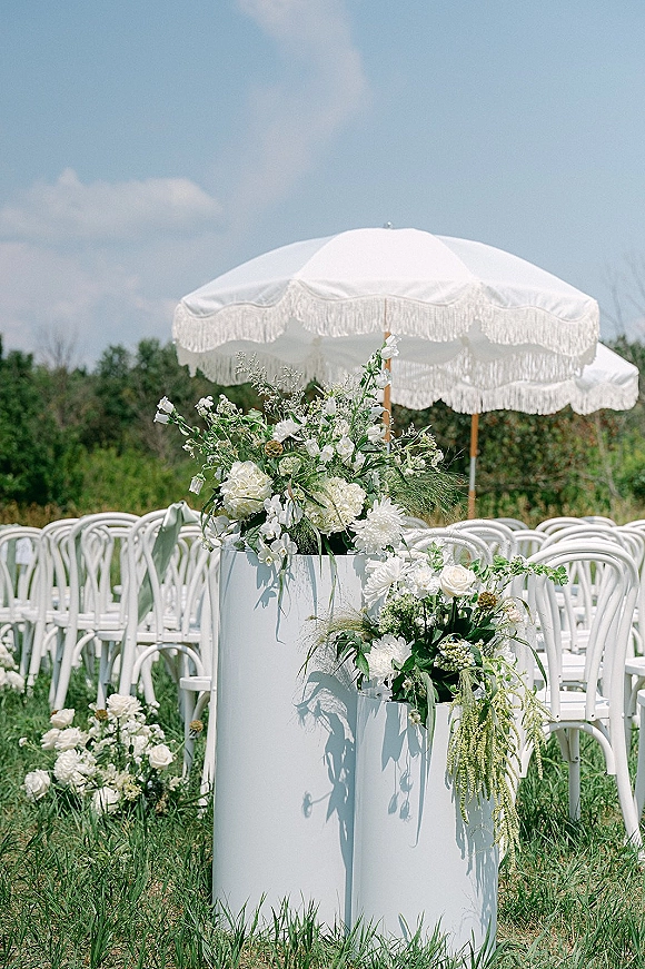 Ceremony aisle decor with white pedestal plinths and outdoor ceremony aisle decor, featuring lush white florals and a fringe parasol on a grassy lawn