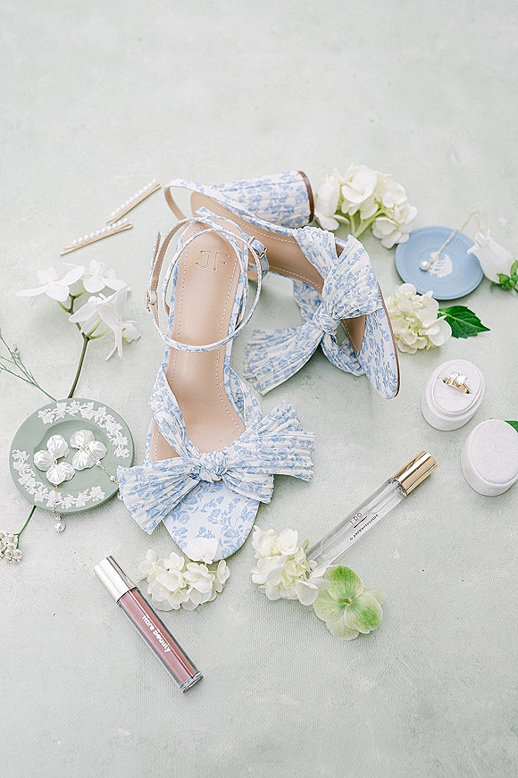 Bridal shoes with blue floral bow heels arranged in a flat lay with wedding rings, pearls, perfume, and hydrangeas on gray fabric
