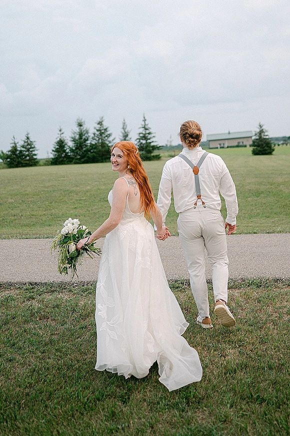 Couple portrait of newlyweds holding hands walking away on a paved path, bride glancing back with bouquet, evergreens and farm building behind
