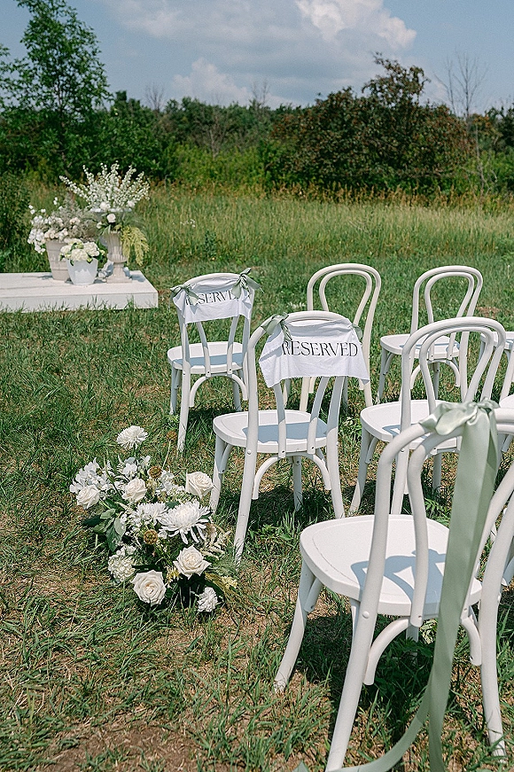 Ceremony seating with outdoor wedding chairs, white bentwood rows with reserved signs and sage ribbons in a grassy meadow under cloudy sky