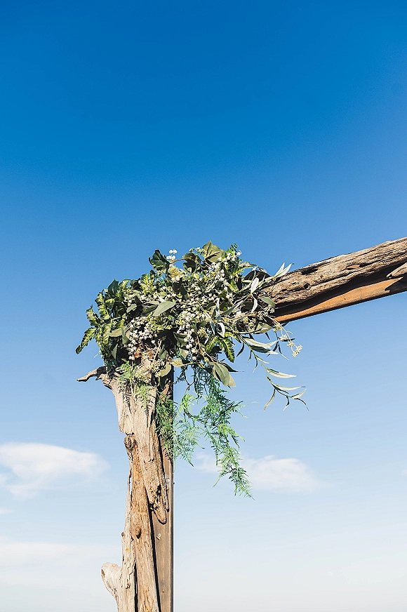 Wedding ceremony arch with a rustic wooden arbor draped in eucalyptus and white flowers, set against a blue sky with clouds