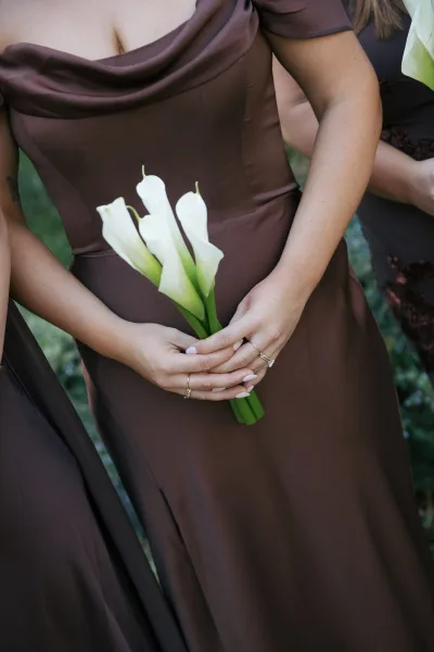 Bridesmaid bouquet, a calla lily bouquet, held by manicured hands, framed by greenery on a grass lawn.