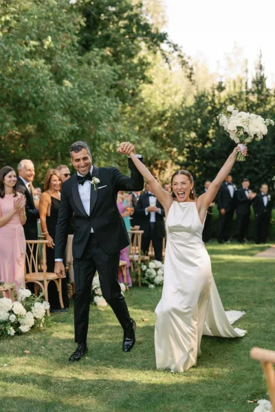 Wedding recessional as bride and groom walking aisle, holding hands while she lifts her bouquet overhead as guests cheer in a garden setting