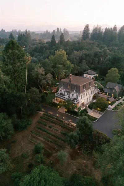 Wedding venue exterior with aerial wedding venue view, glowing string lights over patio umbrellas, set among vineyard rows at dusk