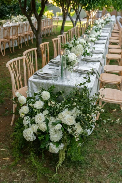 Reception tablescape with an outdoor wedding reception table set with white florals, greenery garland, and taper candles on a garden lawn