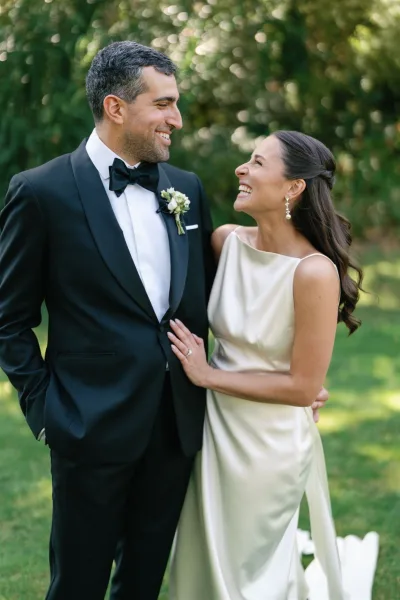 Couple portrait of bride and groom laughing, her hand on his chest, in garden greenery; groom in black tux with boutonniere