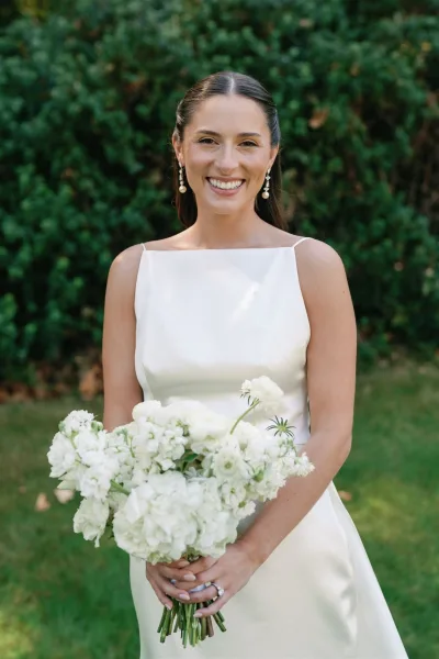 Bridal portrait of a smiling bride holding a white wedding bouquet, wearing pearl drop earrings and ring before green hedges