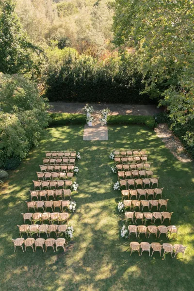 Ceremony setup for an outdoor wedding ceremony with wood chairs flanking an aisle runner, white florals on pedestals on a garden lawn