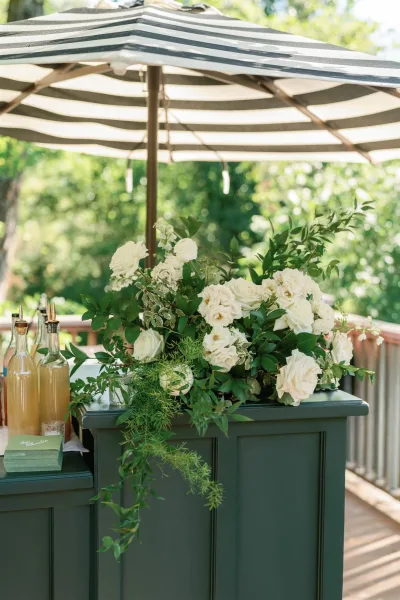 Wedding bar setup with an outdoor wedding bar cabinet, glass bottles and drink dispensers, shaded by a striped umbrella on a garden deck