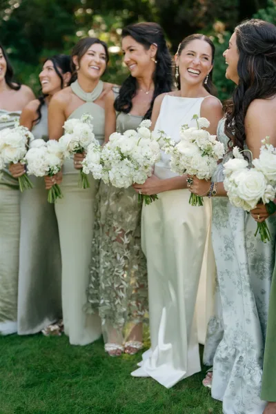 Bridesmaid group photo with bridesmaids holding bouquets of white roses and hydrangeas, laughing in sage satin dresses on a garden lawn