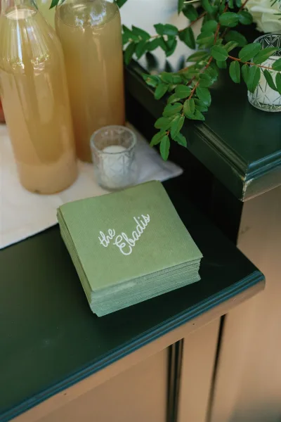 Wedding cocktail napkins and custom wedding napkins in sage green script on a wooden bar with glass bottles, votive candle, and greenery stems