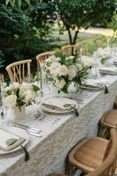 Reception tablescape with white rose centerpieces, greenery, and taper candles on a patterned tablecloth at an outdoor garden lawn dinner setting