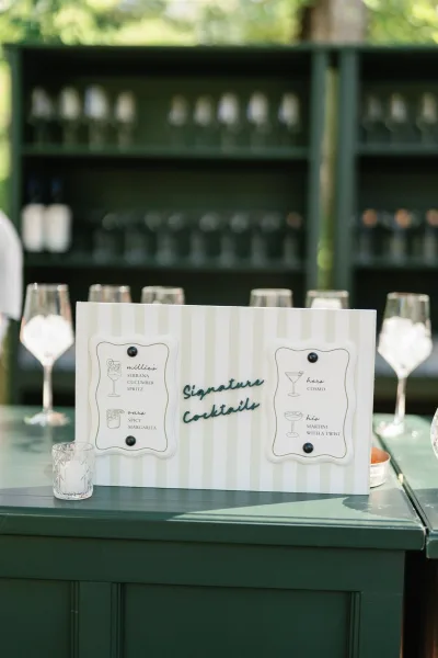 Signature cocktail sign wedding bar sign on a green counter with striped board, menu cards pinned above glasses and bottles in sunlight greenery