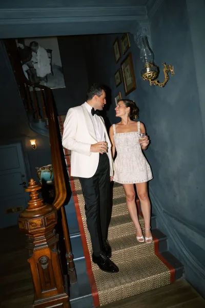 Couple portrait of newlyweds holding cocktail glasses on a staircase, bride in embellished short dress and groom in white tuxedo jacket.
