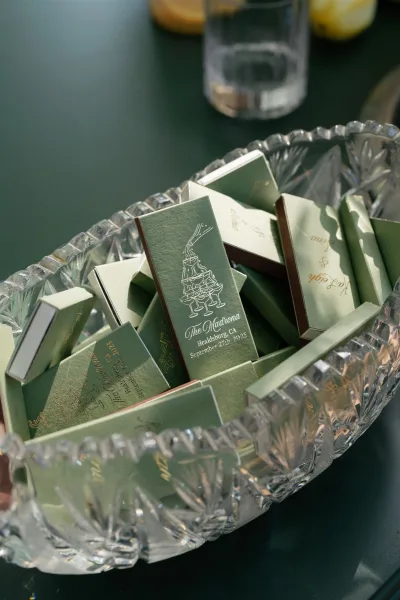 Wedding matchboxes and custom wedding matchboxes in a crystal cut glass bowl with gold foil text on a dark green tabletop beside cocktails