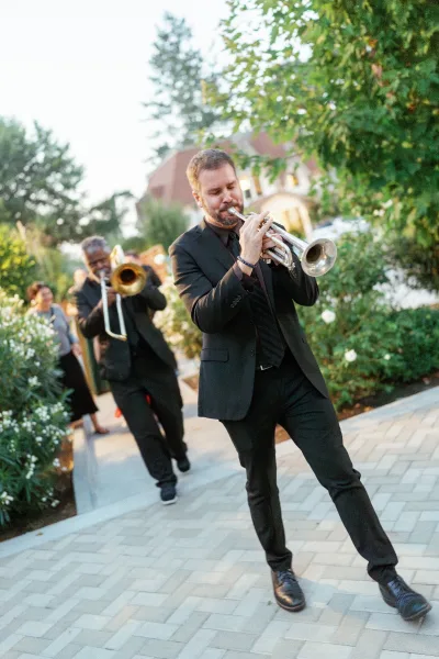 Wedding band in black suits marching with a trumpet down a garden walkway as guests watch near white flowers and a building facade