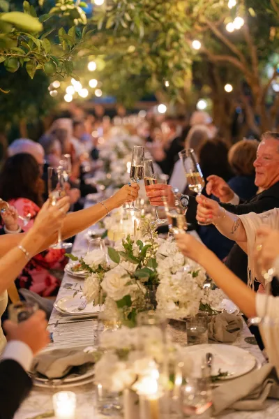 Wedding reception toast as guests raise champagne flutes amid candlelight and white floral centerpieces under garden string lights at night