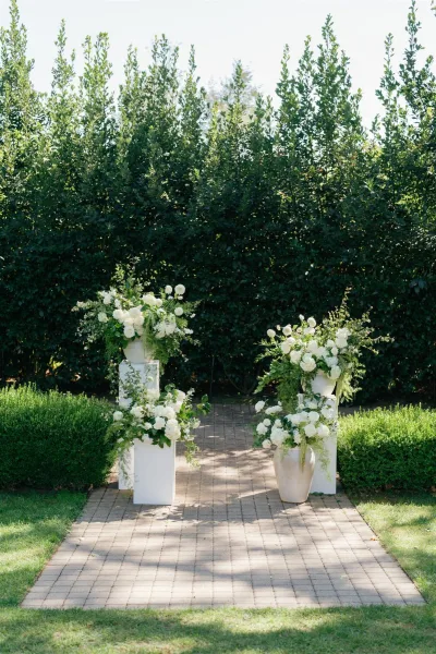 Ceremony aisle decor with white floral arrangements and greenery on pedestal urns lining a brick walkway through manicured garden hedges