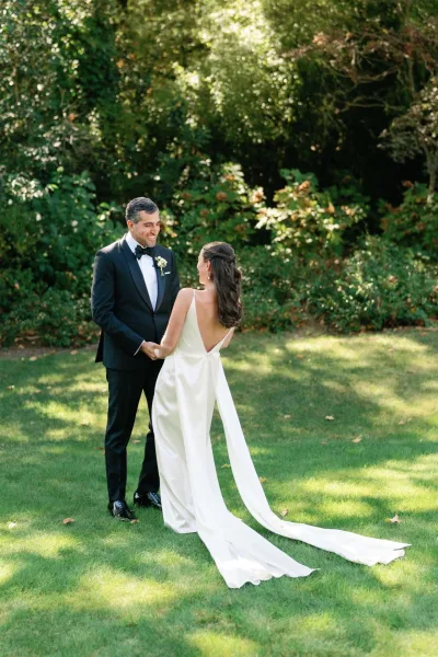 First look moment as bride in a low back gown with long train holds hands with tuxedoed groom on a sunlit garden lawn