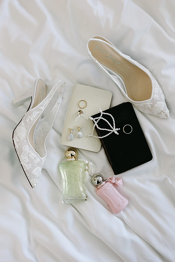 Wedding flat lay of lace bridal shoes, wedding rings, pearl earrings, vow books and perfume bottles arranged on white fabric drape