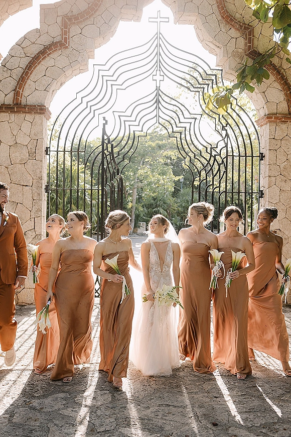 Bridesmaids with bride in rust dresses walk beside a veiled bride holding white calla lily bouquets under a stone archway in sunlight