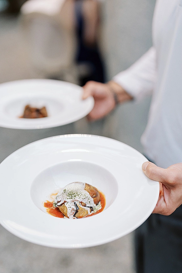 Wedding catering staff serving a plated wedding dinner on white plates with sauce, herb garnish, and blurred guests in an indoor venue