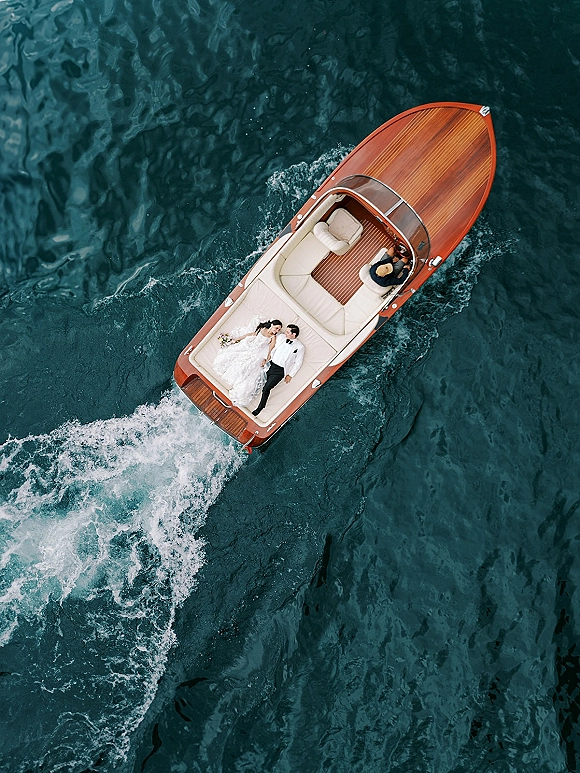 Couple portrait of bride and groom on a wooden speedboat, holding hands with a bouquet, surrounded by open water and wake