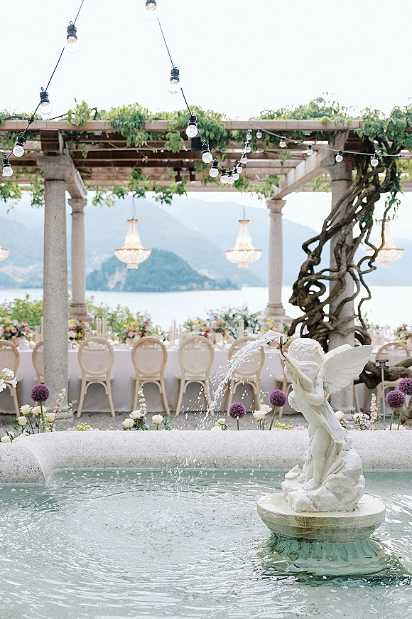Outdoor reception setup with a long banquet table setting under a vine-covered pergola, lit by string lights and chandeliers by the lake and mountains