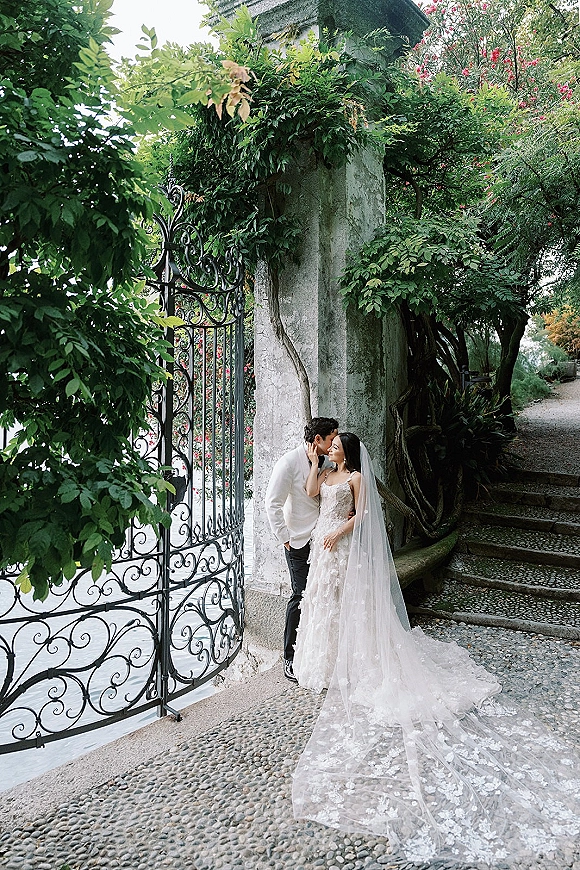 Wedding couple portrait of bride and groom kissing, her long veil draped by a wrought iron gate along garden steps and stone wall