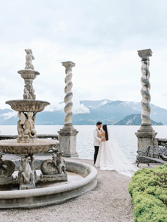 Couple portrait of bride and groom embrace, her long cathedral veil trailing by a stone fountain on a terrace with lake and mountains beyond