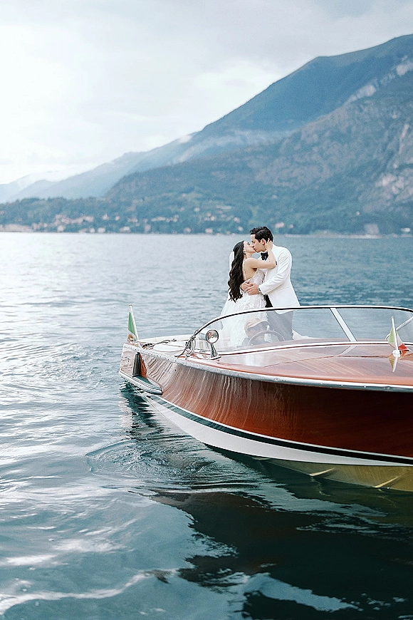 Wedding kiss portrait of bride and groom on boat, her veil blowing as he wears a white jacket, with lake and mountains behind under clouds