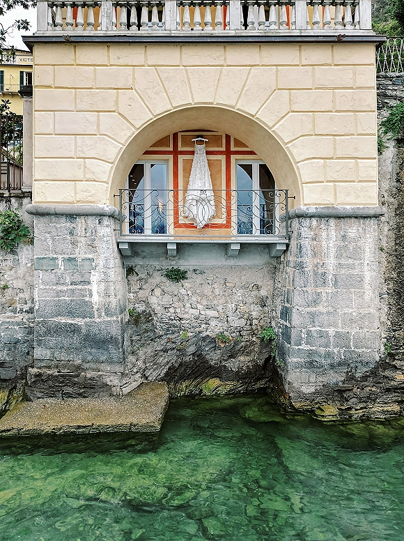 Wedding dress, lace wedding dress hanging from a balcony railing by glass doors, with arched stone facade and waterfront behind