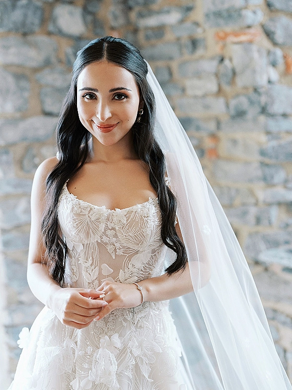 Bridal portrait of a bride in a lace wedding dress and cathedral veil, smiling with drop earrings, posed against a stone wall
