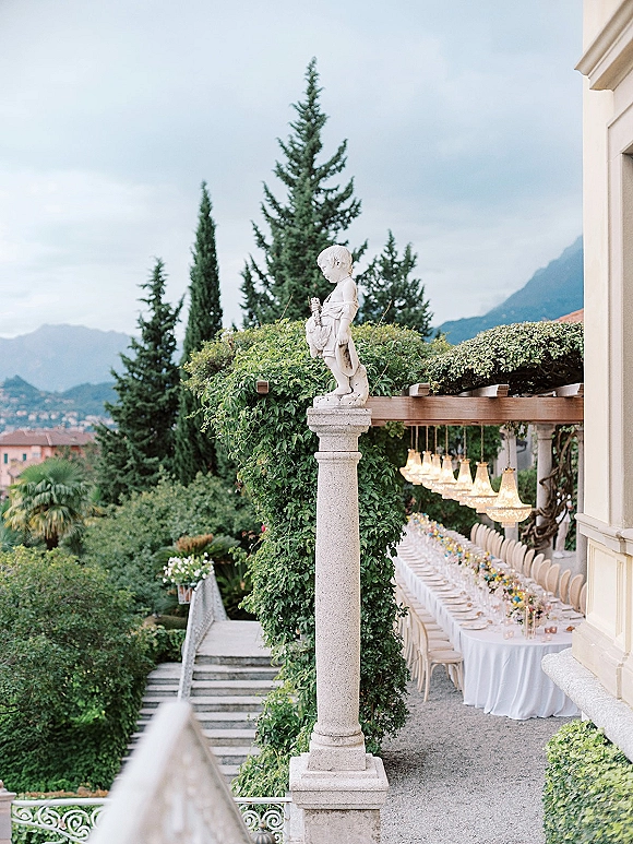 Outdoor reception tablescape with a long banquet table wedding setup, white linens, floral garland and taper candles under chandeliers on a villa terrace with mountain views