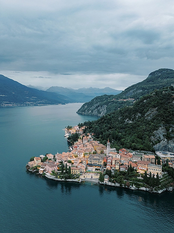Lake town aerial view of a lakeside village panorama with terracotta rooftops, church steeple, and waterfront buildings by mountains under clouds
