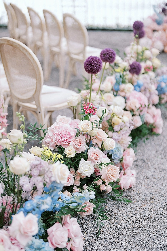 Ceremony aisle florals with roses, hydrangea and allium nestled in greenery along a gravel patio aisle beside ivory oval-back chairs